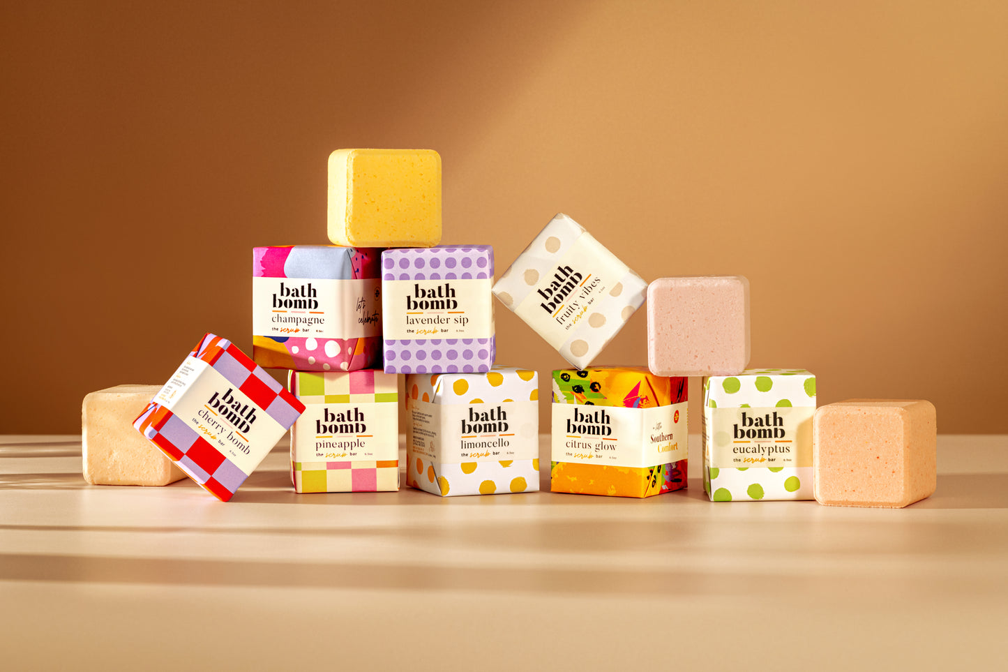 A lineup of natural bath bombs from The Scrub Bar displayed with bold, colorful gift-ready packaging. Each square bomb showcases our luxury bath oil blends, bubble bath bubbles, and skin-loving bath bomb ingredients—all stacked to show off their handcrafted appeal.