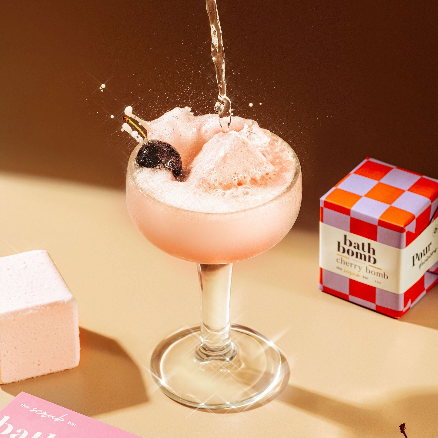 The Scrub Bar’s Cherry Mimosa bath bomb fizzes in a wine glass with foam, cherries, and luxe pink packaging nearby