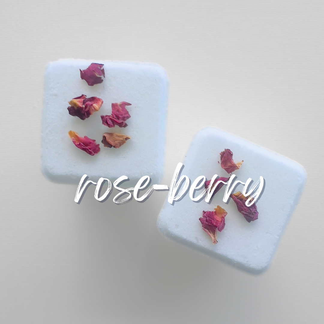 two square-shaped bath bombs from The Scrub Bar's Rose Sangria collection. Each bath bomb is topped dried rose petals. The bombs are set against a plain, light background that emphasizes their pristine white color and the vibrant color of the rose petals.. Across the photo with the word rose-berry