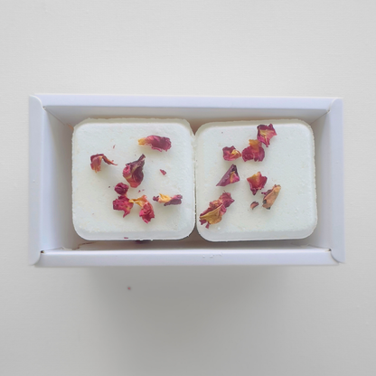 two square-shaped bath bombs from The Scrub Bar's Rose Sangria collection. Each bath bomb is decorated with scattered dried rose petals, enhancing their delicate and romantic appeal. The bombs are set against a plain, light background that emphasizes their pristine white color and the vibrant contrast of the rose petals.