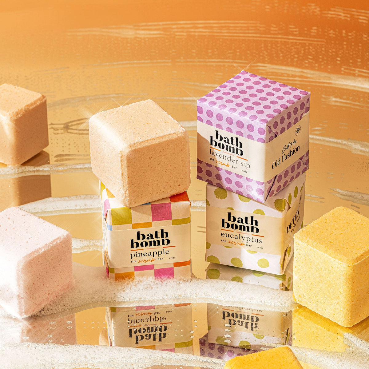 The Scrub Bar's Natural bath bombs  wrapped shown across a wet, reflective surface, surrounded by foam and bubble bath bubbles. Highlighting the foamy fizz and artisan look.